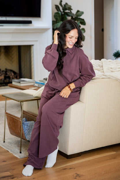 A woman wearing a matching mauve-purple lounge set consisting of a long-sleeve top and wide-leg pants, sitting on a beige sofa in a living room.
