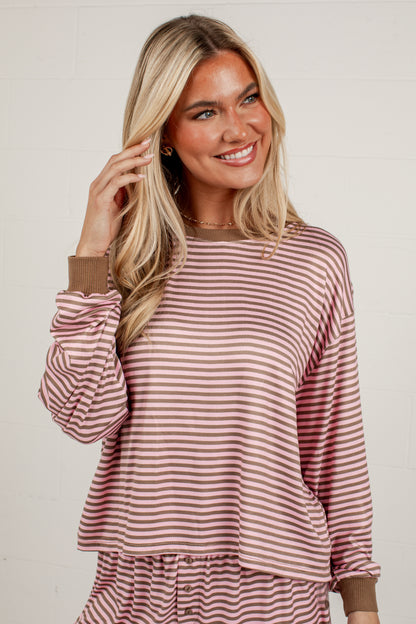 A smiling blonde woman wearing a long-sleeve dusty rose and white striped top with brown ribbed cuffs, standing against a light off-white brick wall.