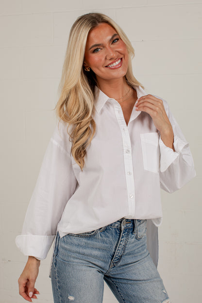 A smiling blonde woman wearing a white long-sleeve button-up shirt with rolled cuffs and light-wash blue jeans, standing against a light off-white brick wall.