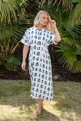 Woman in a white dress with blue floral pattern standing outdoors with greenery in the background