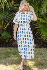 Woman in a white dress with blue floral pattern standing in front of green foliage.