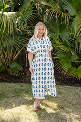 Woman in a white dress with blue floral pattern standing in front of green foliage.
