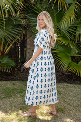 Woman in a white dress with blue floral pattern standing outdoors with greenery in the background