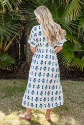 Woman wearing a white dress with blue floral pattern standing in a garden.