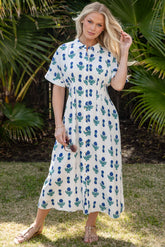 Woman in a white dress with blue floral pattern standing in front of tropical plants.