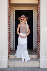 A woman stands on the tiled steps of a doorway wearing a beige sleeveless top, a white textured skirt, a wide-brimmed beige hat, and white pointed shoes.