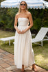 Woman in a strapless white dress standing under a striped umbrella in a garden setting.