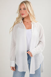 Woman wearing a white sheer blouse over a white top with blue jeans on a light gray background