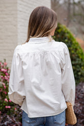 Woman wearing a light-colored blouse and blue jeans, standing outdoors with blurred background
