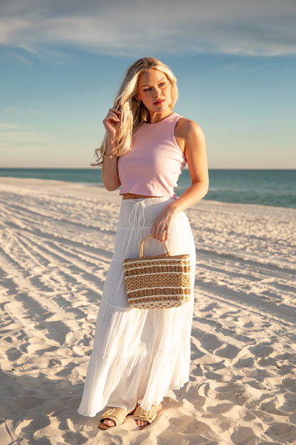 Woman on a beach holding a woven bag, wearing a pink top and white skirt.