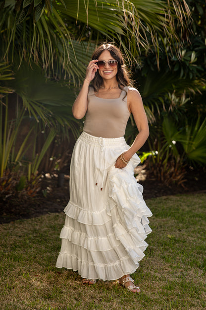 Woman in beige top and white ruffled skirt standing outdoors with greenery in the background