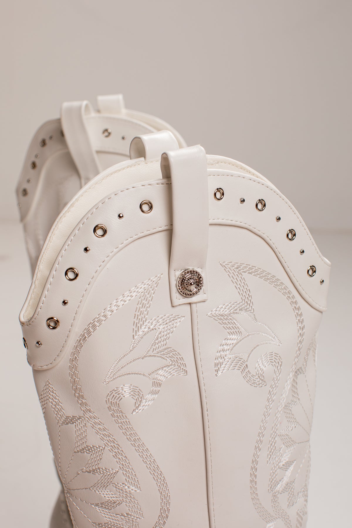 Close-up of a white western boot with silver-toned studs, decorative stitching, and a central concho button, shown against a neutral light gray background.
