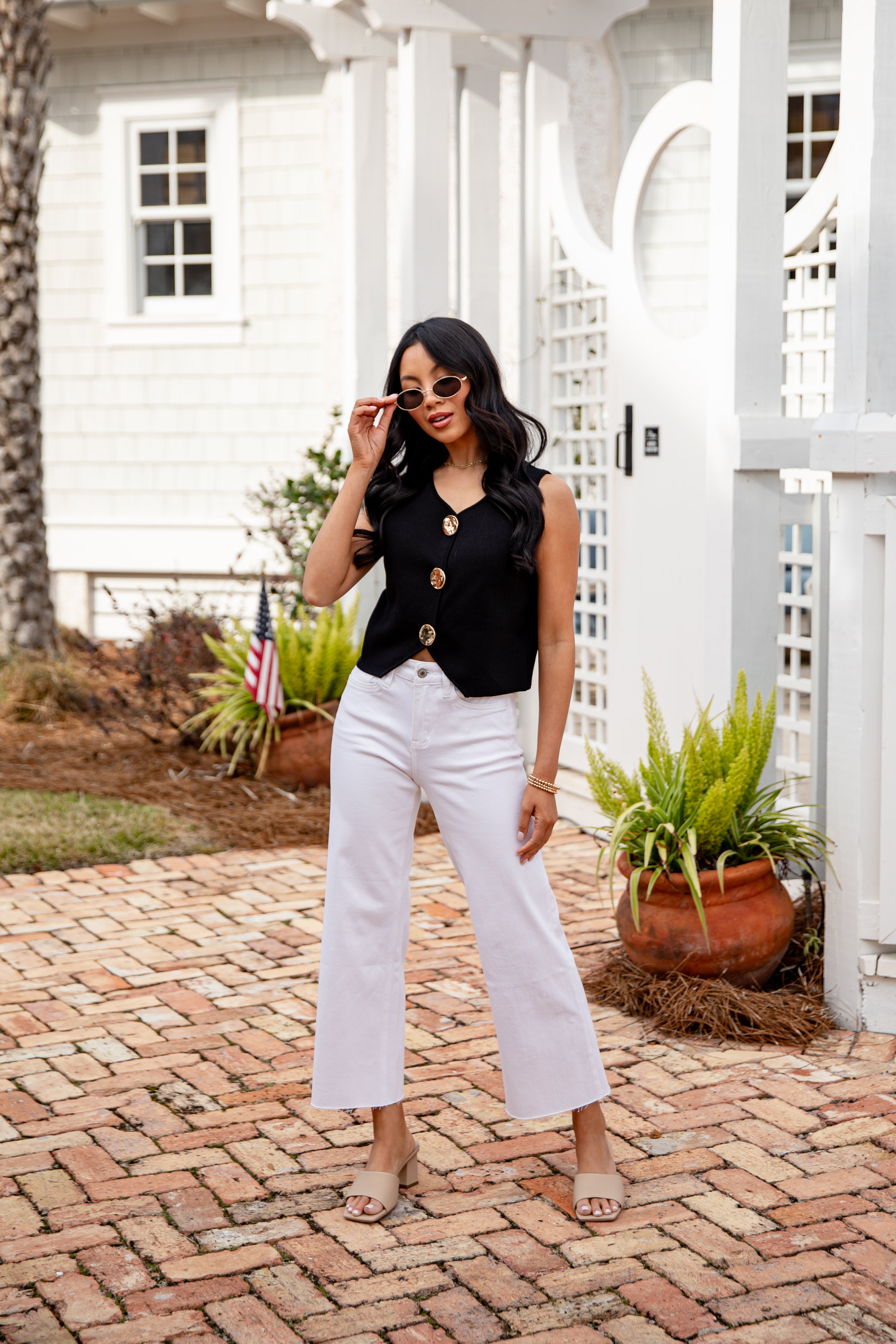 Woman in black top and white pants standing on a brick patio with plants and a white house in the background.