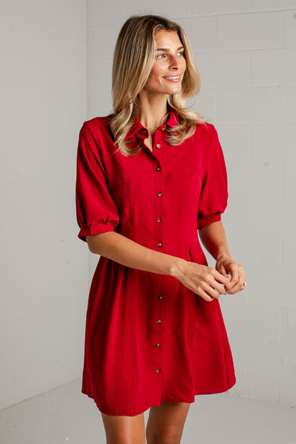 A woman wearing a bright red short-sleeve button-front dress with a pointed collar and tie detail at the neck, standing against a light gray brick wall.