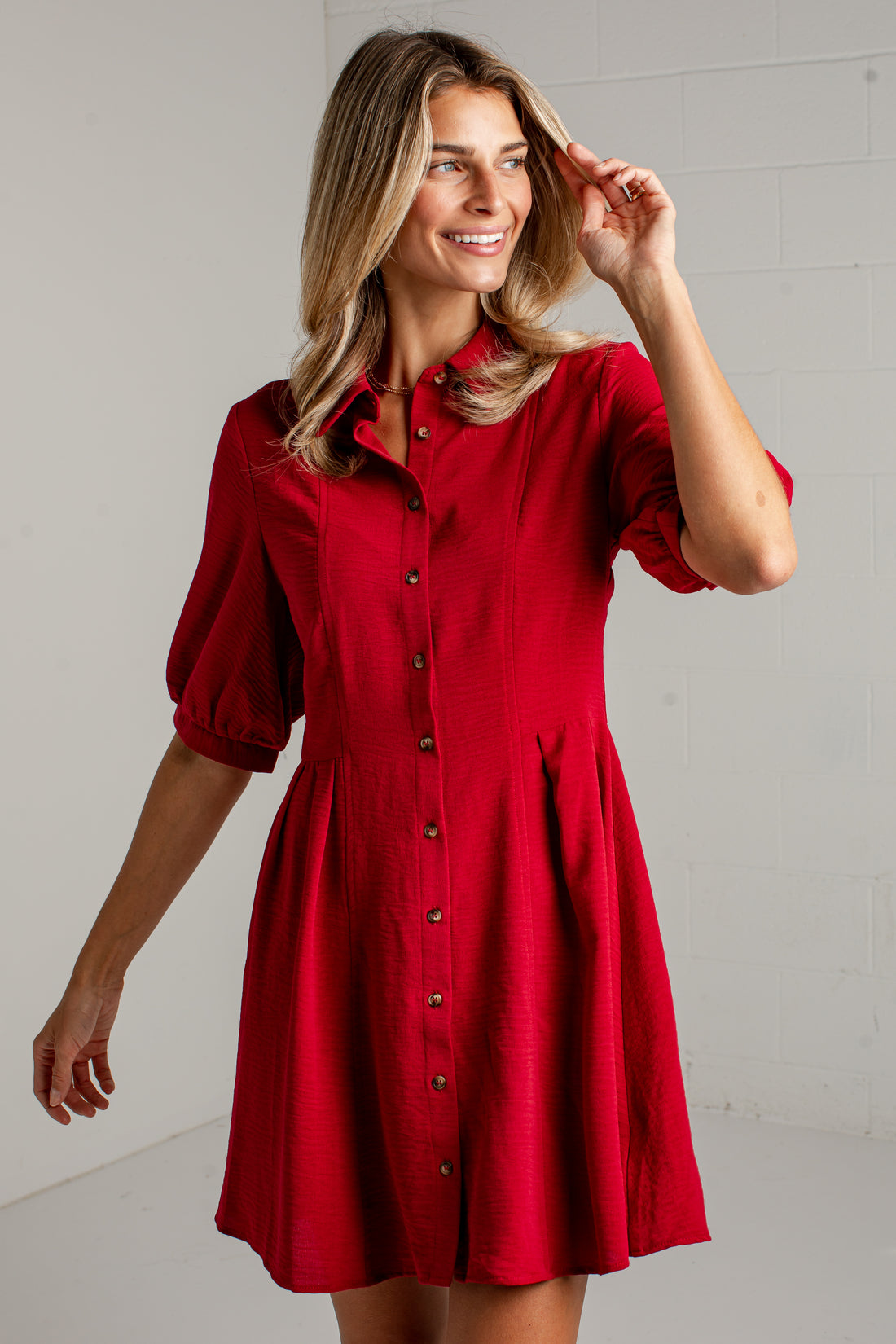A woman modeling a red short-sleeve button-up dress with a flared skirt, photographed against a light gray brick wall.