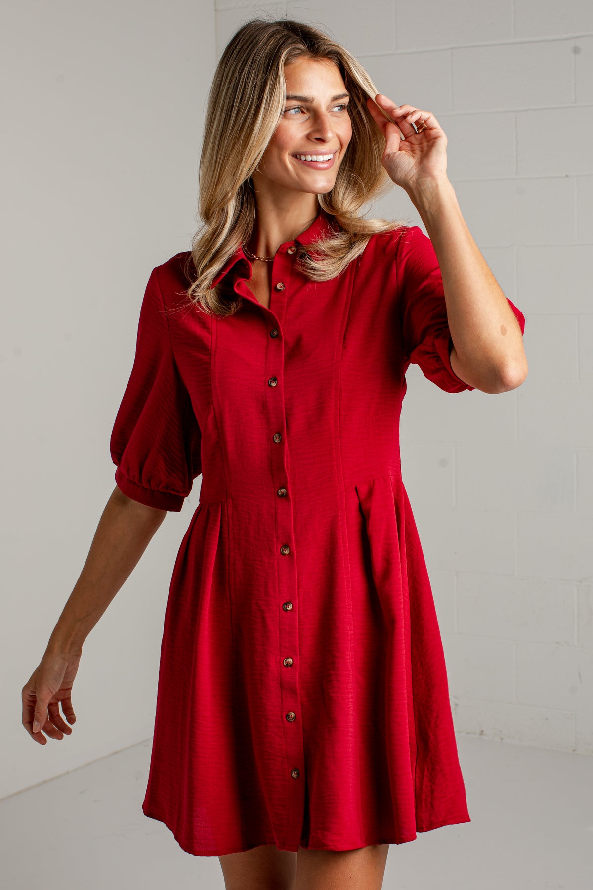 A woman modeling a red short-sleeve button-up dress with a flared skirt, photographed against a light gray brick wall.