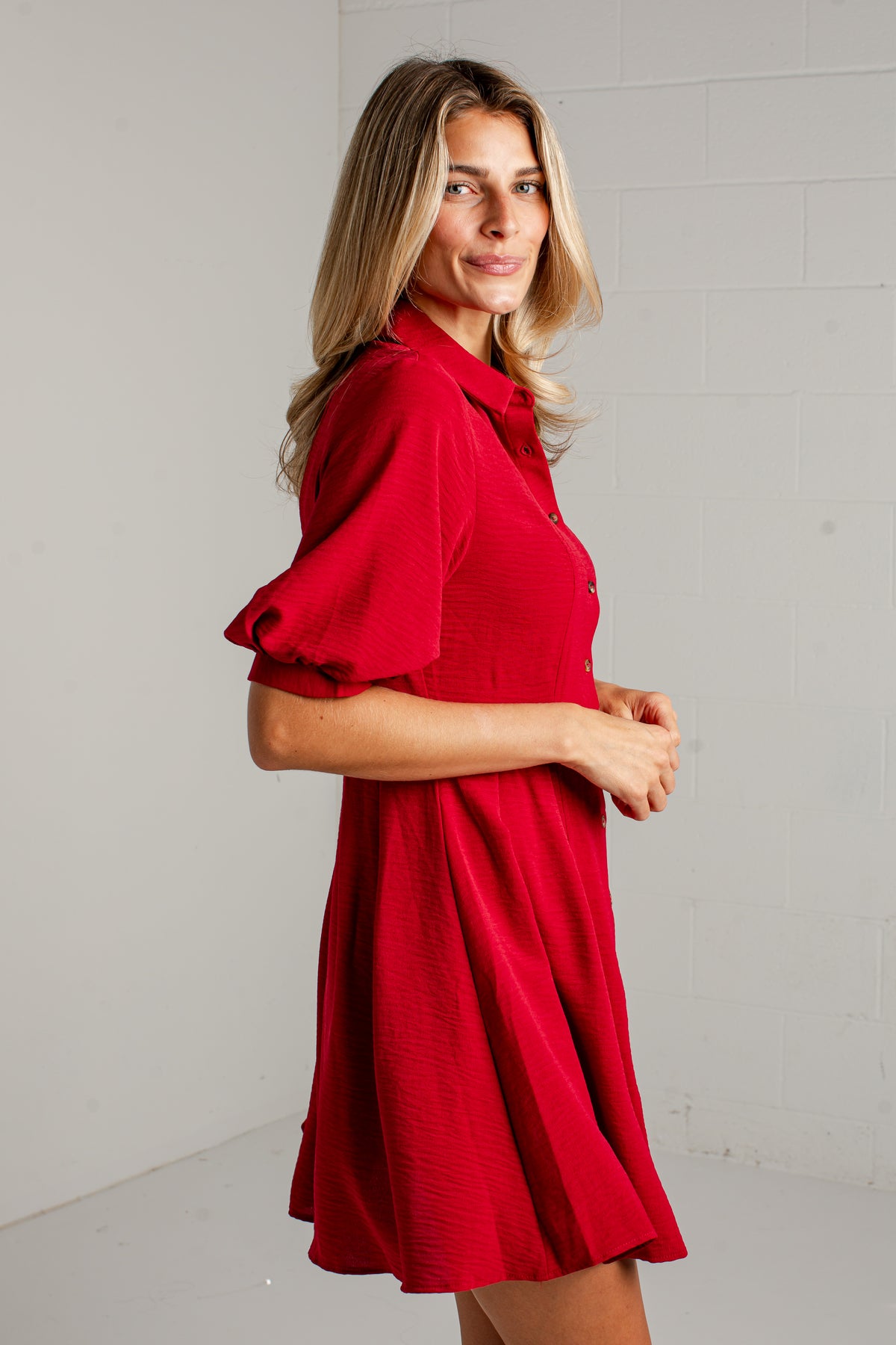 A woman modeling a short-sleeve red dress with a collar and button front, standing against a light gray brick wall.