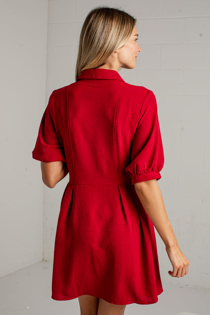 A woman wearing a bright red short-sleeve dress, shown from the back as she stands against a white brick wall.