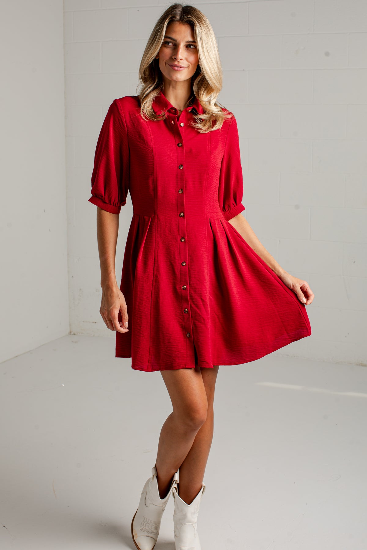 A woman modeling a bright red short-sleeve button-front dress with a flared skirt, paired with white cowboy boots, standing in a white studio corner.