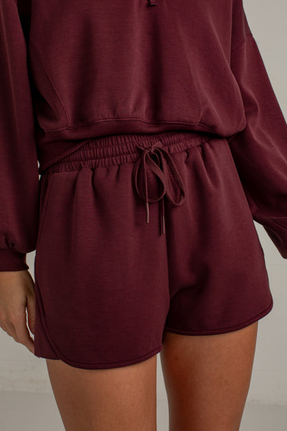 A close-up of a person wearing a matching burgundy lounge set, featuring a cropped sweatshirt and drawstring shorts, against a neutral light gray background.