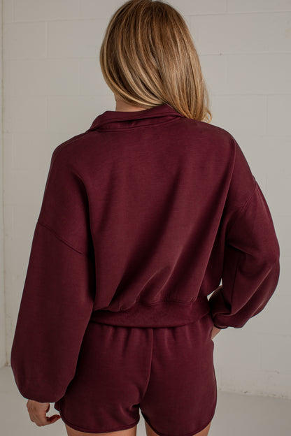 A woman viewed from behind wearing a matching burgundy lounge set consisting of a cropped long-sleeve sweatshirt and shorts, set against a light gray brick wall.