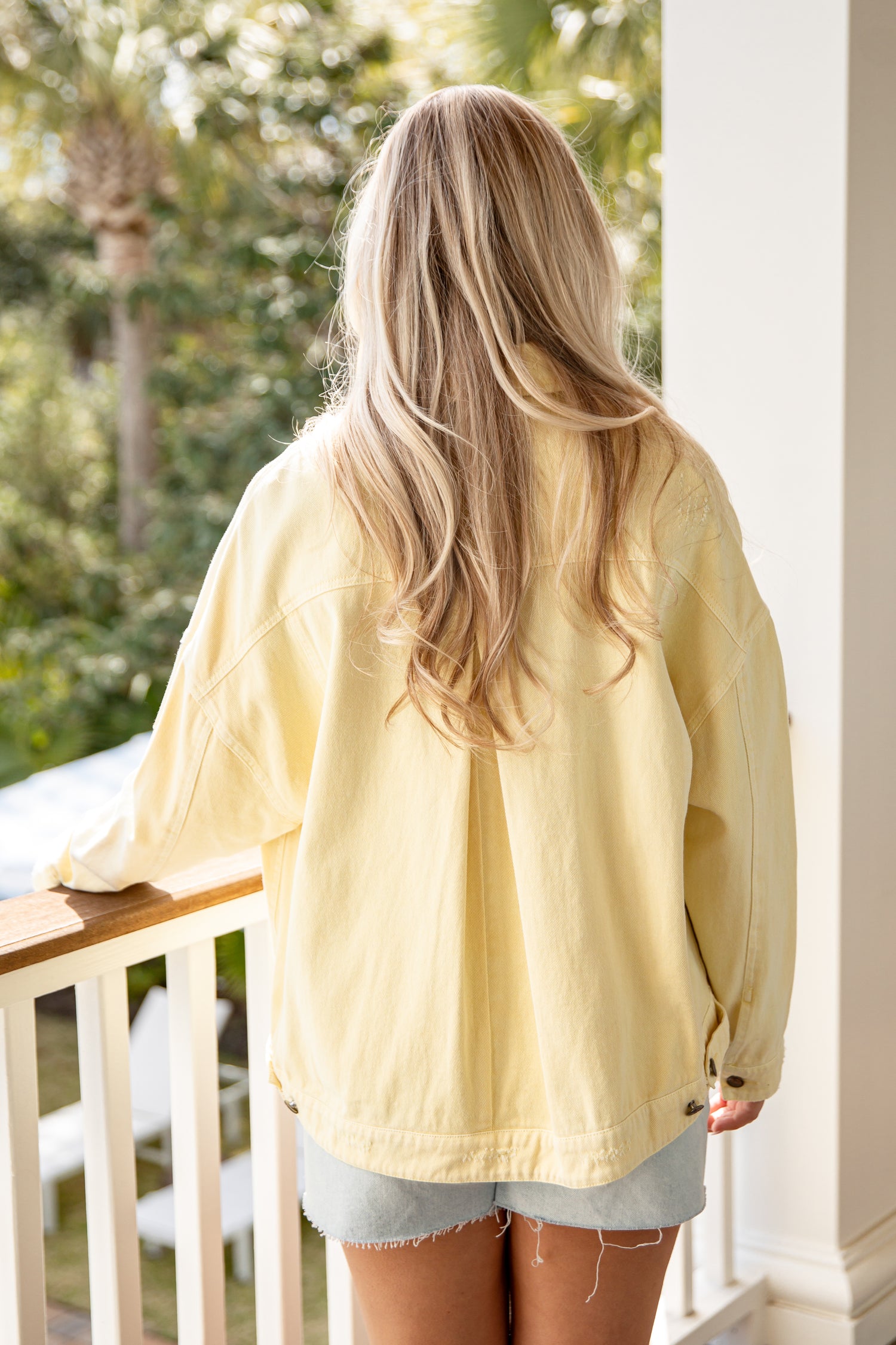 Person wearing a yellow jacket standing on a porch with greenery in the background
