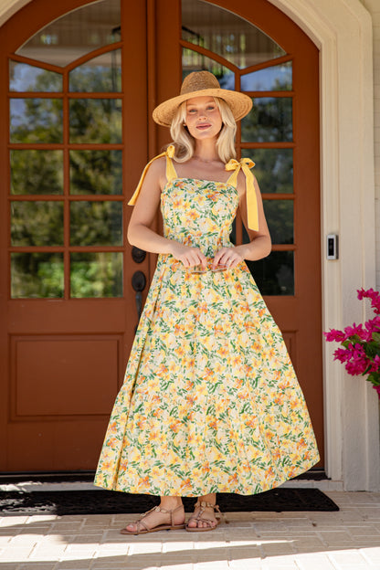 Woman in a floral dress and sun hat standing in front of a brown door with glass panels.