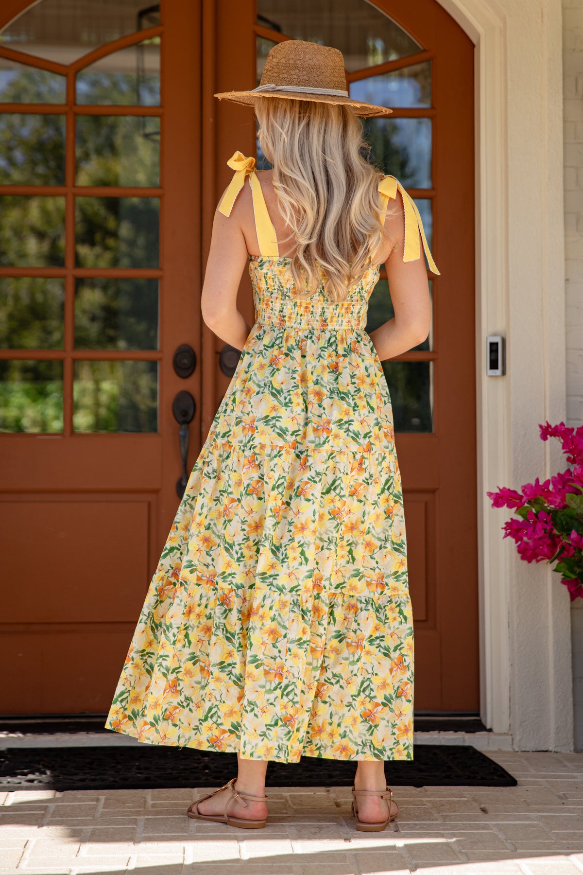 Woman in a floral dress standing in front of a brown door with a pink flower arrangement.
