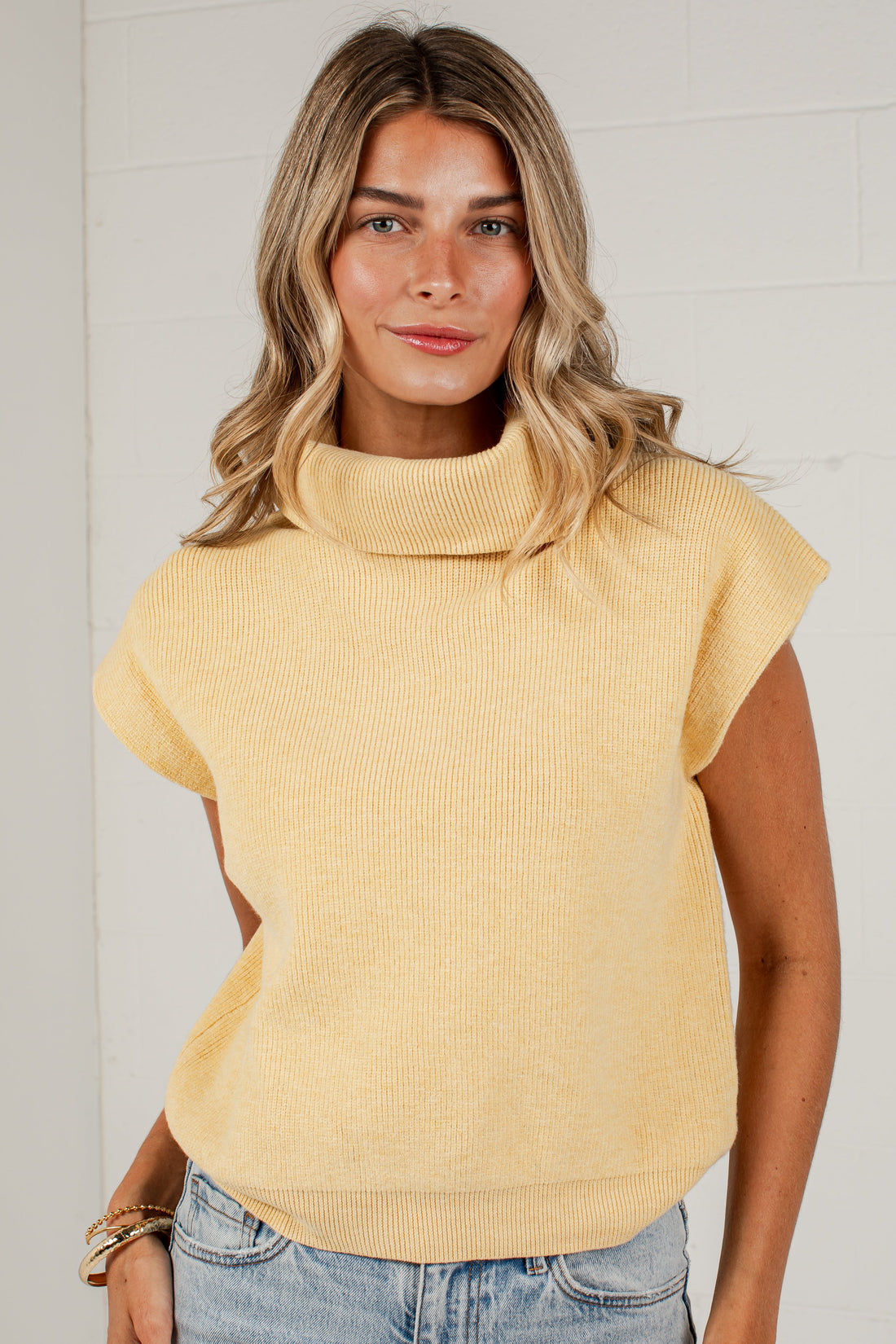 A woman wearing a pale yellow ribbed knit mock neck top with cap sleeves and light blue jeans, standing against a white brick wall.