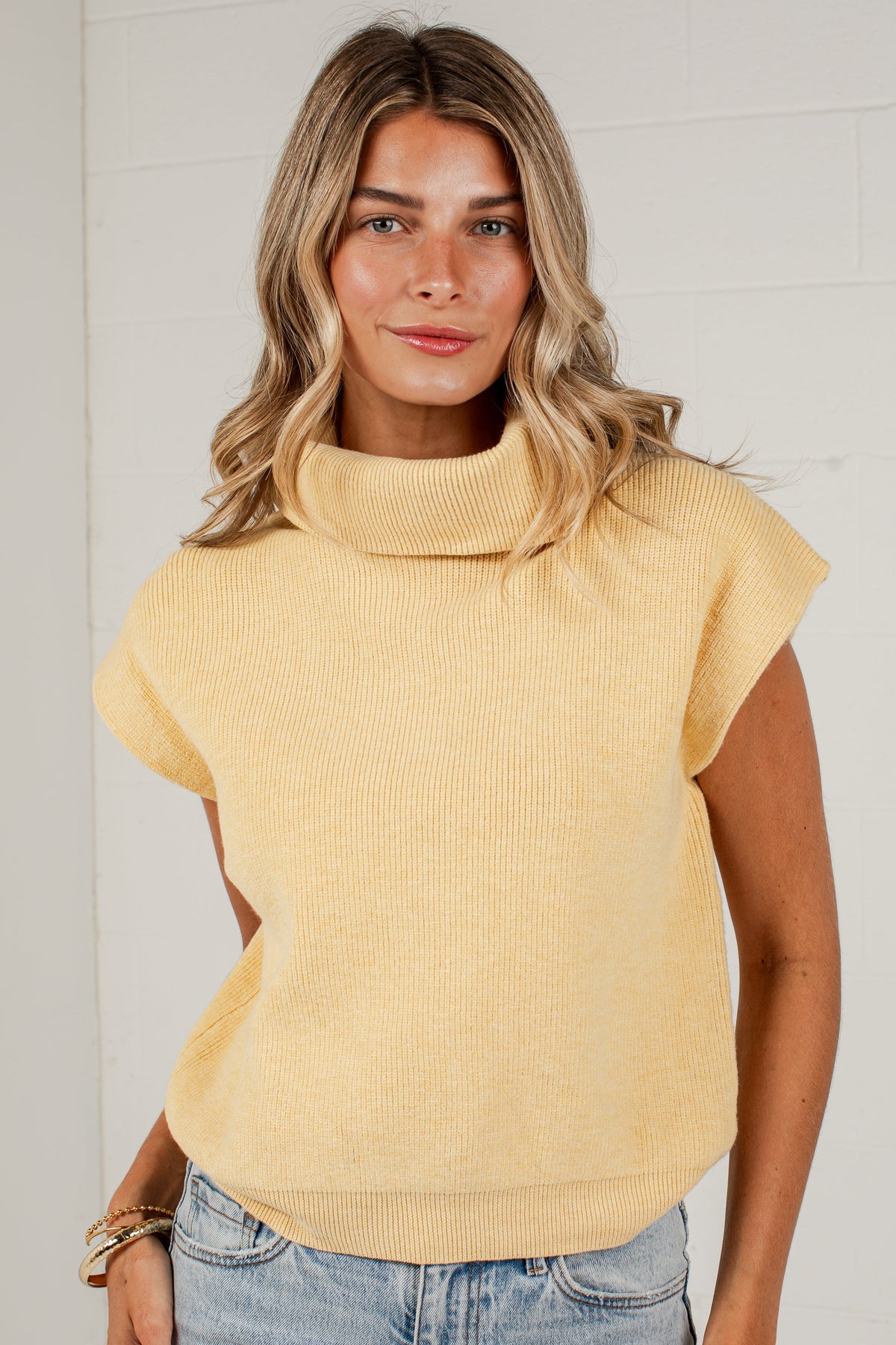 A woman wearing a pale yellow ribbed knit mock neck top with cap sleeves and light blue jeans, standing against a white brick wall.