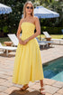 Woman in a yellow strapless dress standing by a pool with lounge chairs and umbrellas in the background.
